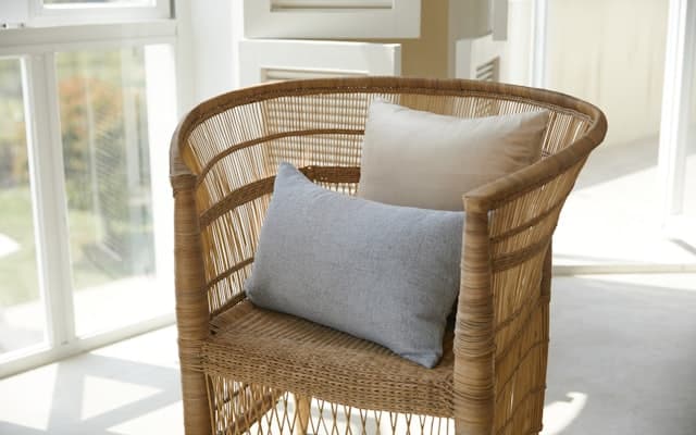 A woven cane lounge chair with two cushions, beside a sunlit window.